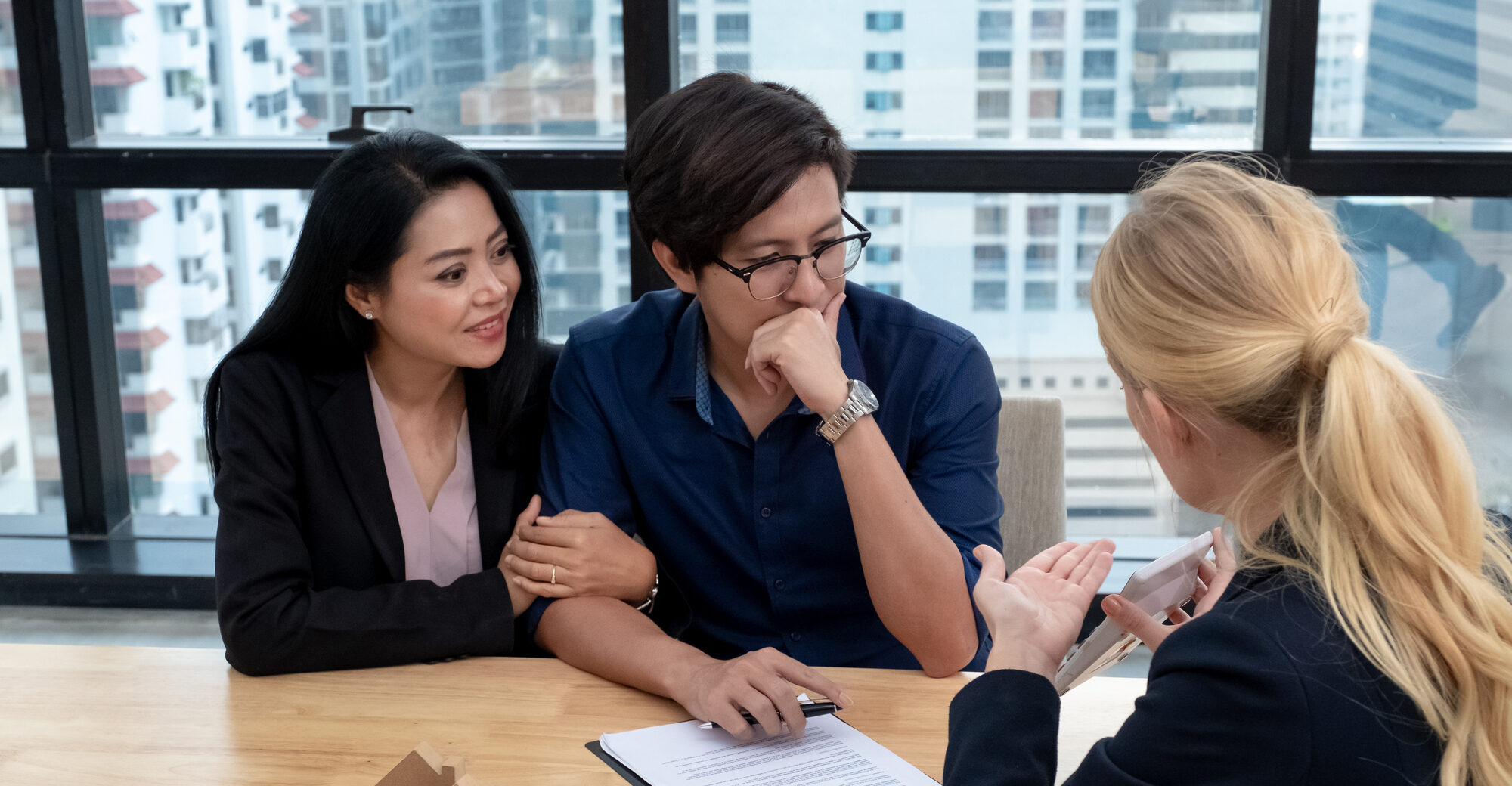 A couple listens attentively to a consultant in a modern office, discussing important documents on a table, with a city view in the background.